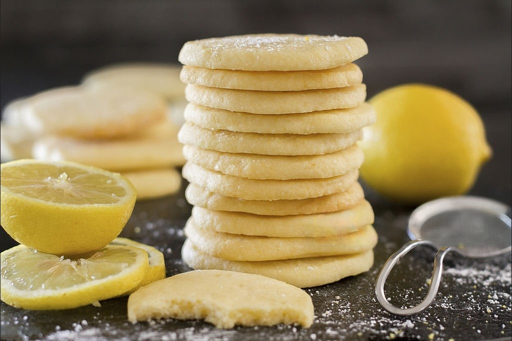Stack of homemade lemon sugar cookies dusted with fine sugar, accompanied by fresh lemon halves and a sifter, evoking a zesty and sweet flavor experience.