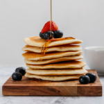 Stack of McDonald's pancakes topped with fresh strawberries and blueberries, drizzled with golden syrup on a wooden serving board, capturing the appeal of a warm and delicious breakfast.