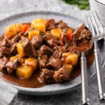 Succulent beef tips crock pot recipe with potatoes and carrots in a rich brown gravy served on a gray ceramic plate with a fork and spoon on the side.