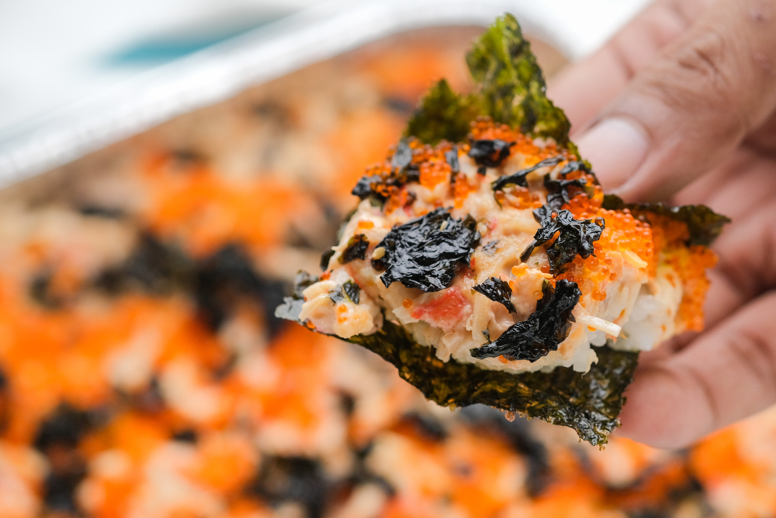 Close-up of a hand holding a piece of bake salmon sushi topped with fish roe and seaweed.