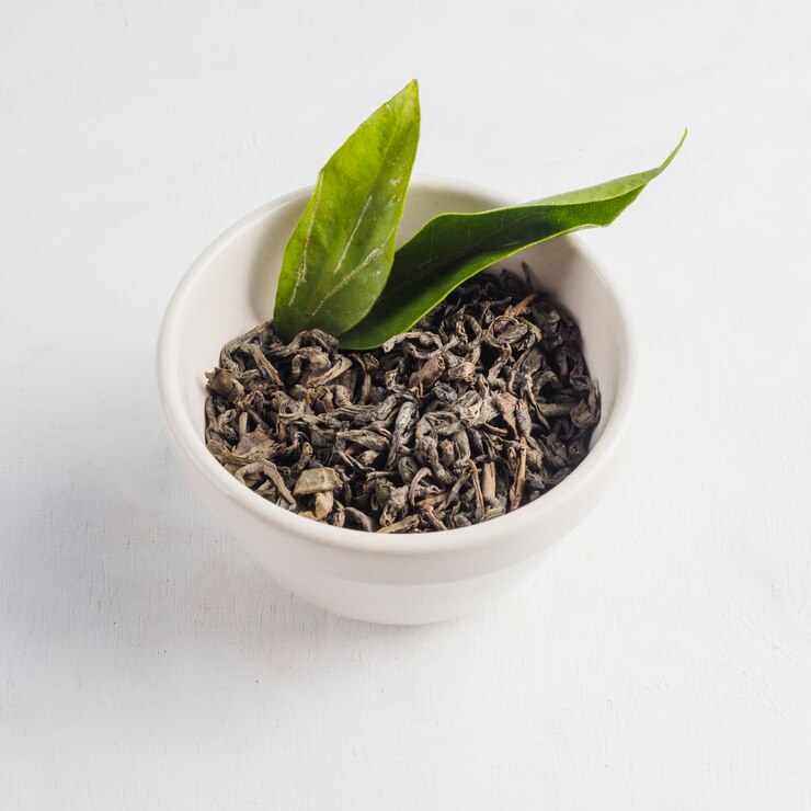 A bowl of loose white tea leaves with fresh green tea leaves on a white background.
