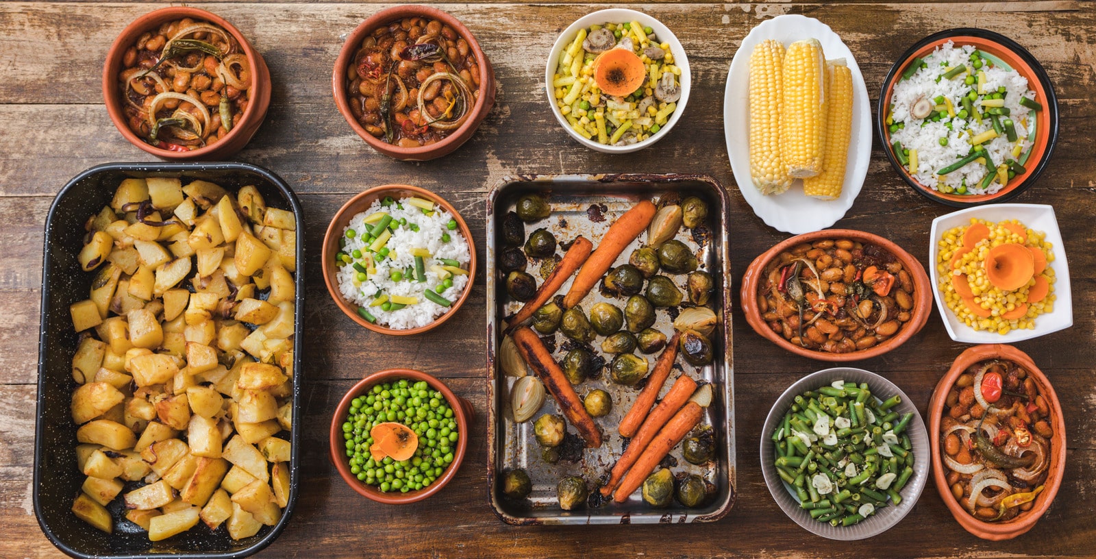 A variety of breakfast casserole side dishes displayed on a wooden table, including roasted potatoes, steamed vegetables, baked beans in earthenware bowls, roasted carrots with brussels sprouts, and bowls of white rice with green peas.
