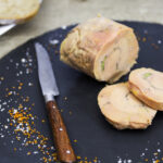 Sliced foie gras terrine on a slate serving board with a rustic wooden-handled knife on the side, scattered with orange and white fleur de sel.