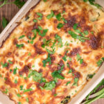 Million dollar chicken casserole topped with melted cheese and fresh parsley, presented in a rectangular ceramic dish, with raw spinach leaves beside it.