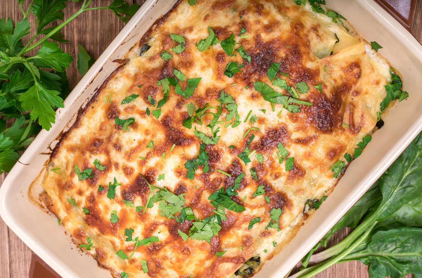 Million dollar chicken casserole topped with melted cheese and fresh parsley, presented in a rectangular ceramic dish, with raw spinach leaves beside it.
