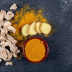 A selection of ginger roots, sliced ginger, and a bowl of turmeric powder on a dark slate background.
