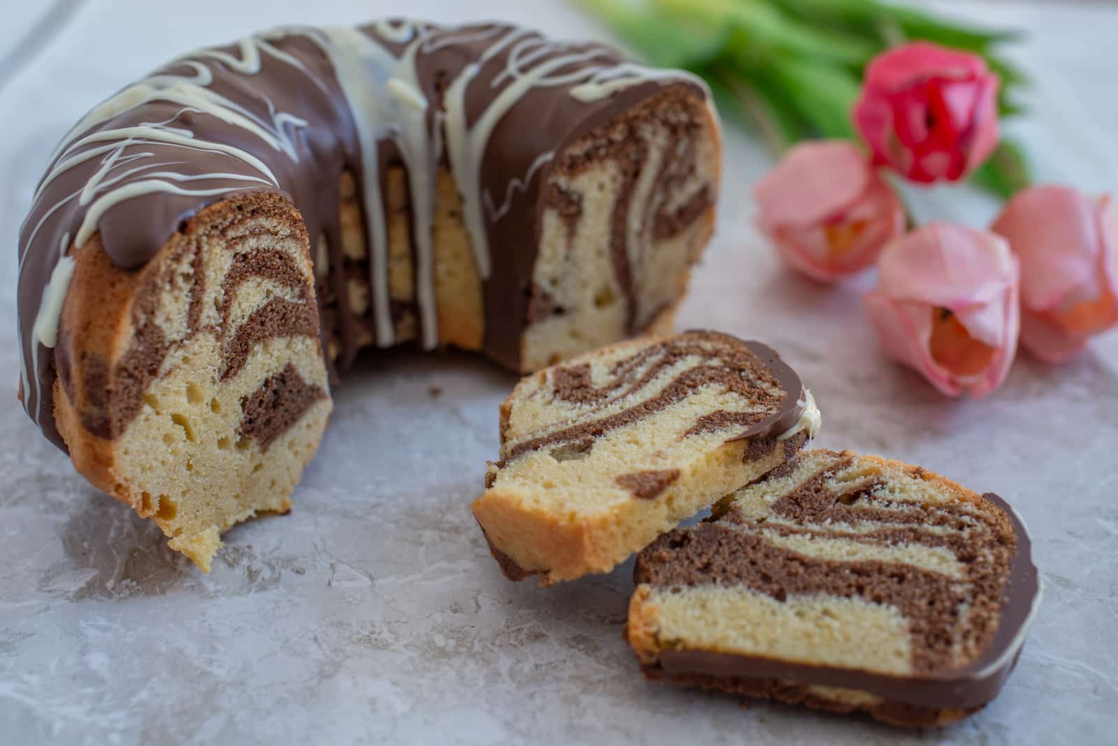 A zebra cake with alternating dark chocolate and vanilla swirl layers, topped with milk and white chocolate drizzle, displayed alongside pink tulips on a marble countertop.
